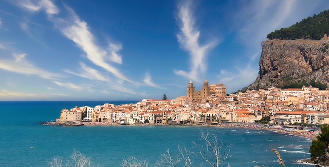 Sicily travel guide image of Cefalu with turquoise water, coastal buildings, mountains, and blue sky